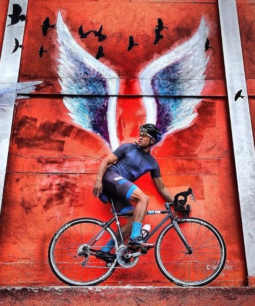 cycliste se tenant sur son vélo devant un mur sur lequel un grafiti en forme d'ailes est peint